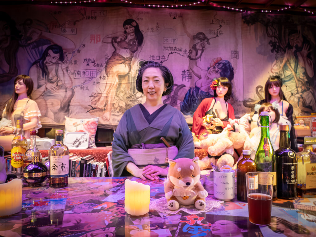 Konta Atsuko in a kimono stands behind a bar of liquor bottles, with love dolls and a painted mural behind her