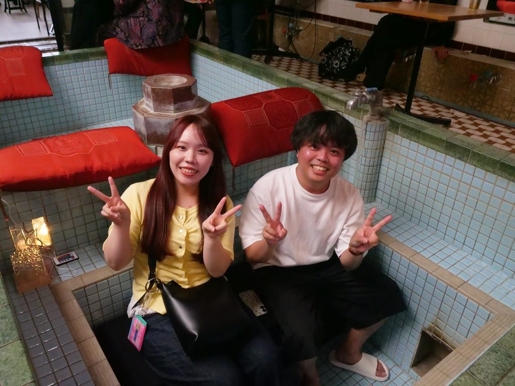 Two audience members sit inside an empty tiled bathtub lined with red cushions, making peace signs at the camera.