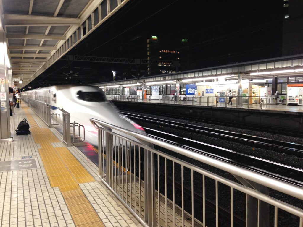 A white and blue Tokaido Shinkansen train stopped at a station platform at night, with the lit concourse and kiosks of the opposite platform visible across the tracks.