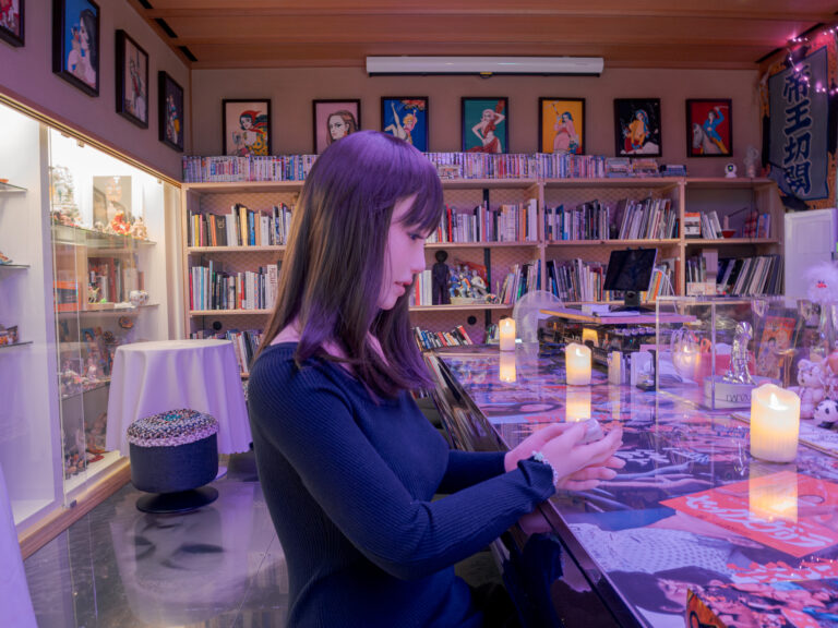 A love doll in a navy long-sleeved dress seated in profile at a glass-topped bar, bookshelves and framed pin-up art behind