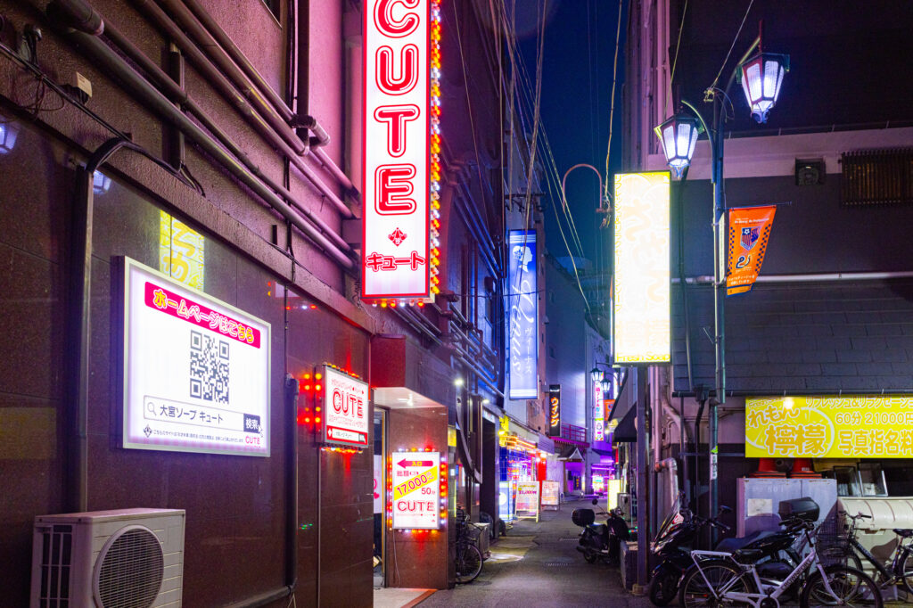A nighttime alley lined with neon signage for soaplands, including one reading “CUTE,” with posted pricing and parked bikes.