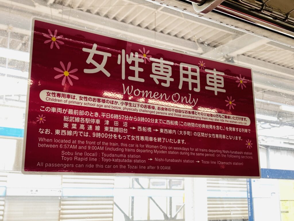 A pink and white 女性専用車 "Women Only" sign above a station platform, with Japanese and English text setting out which trains and hours the women-only car applies to.