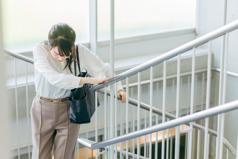 A woman in business attire grips a stairwell railing, leaning forward with her head lowered