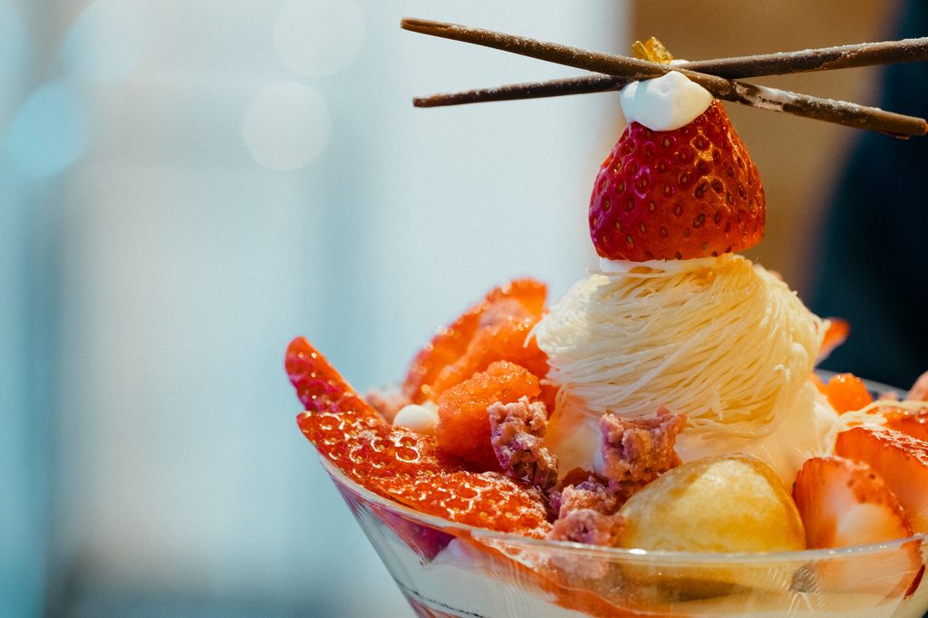 Close-up of a parfait topped with a strawberry, mont blanc cream, and two chocolate sticks crossed above it.