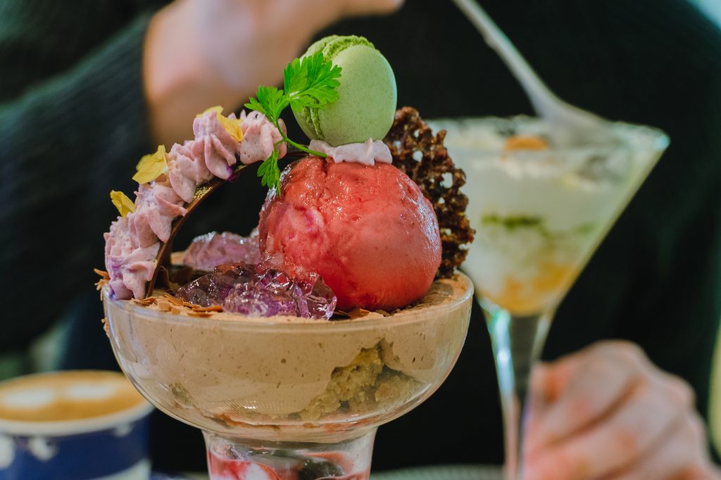 A parfait topped with red sorbet, a green macaron, and berry cream, with a second parfait and a coffee behind it.