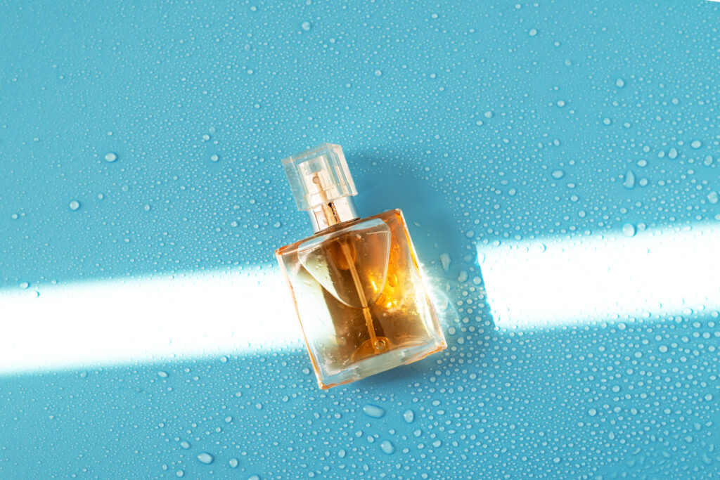 Amber glass perfume bottle lying on a blue surface covered in water droplets, lit by a hard beam of light.