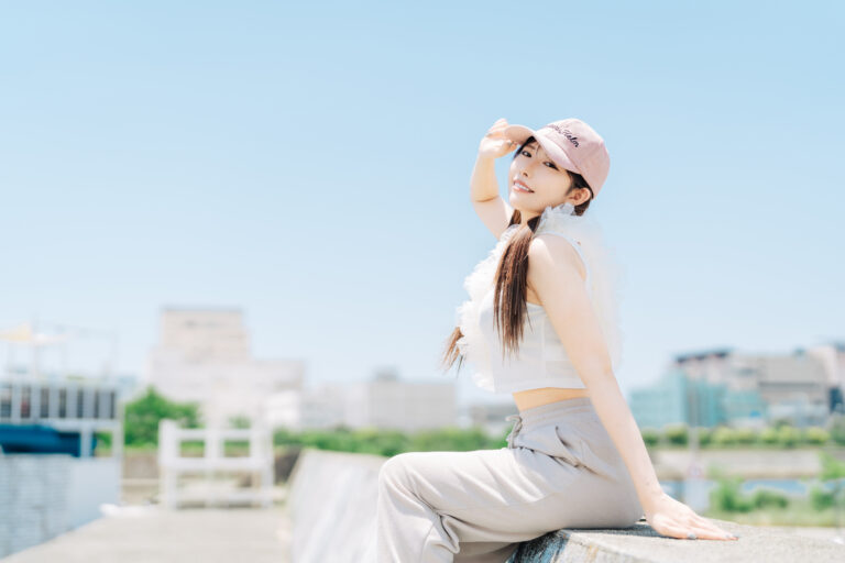 Young woman in a white cropped top, gray sweatpants and a pink cap sits on a waterfront ledge under a clear sky.