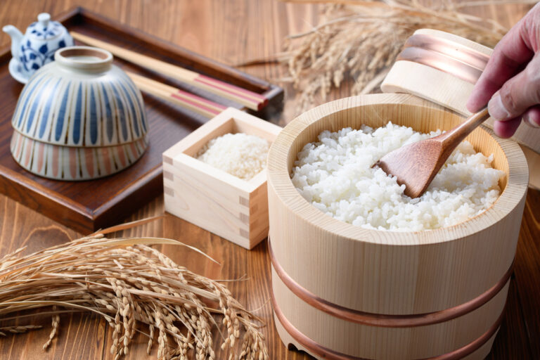 Cooked white rice in a wooden ohitsu with a paddle, beside rice bowls, a masu of uncooked rice and dried rice stalks.