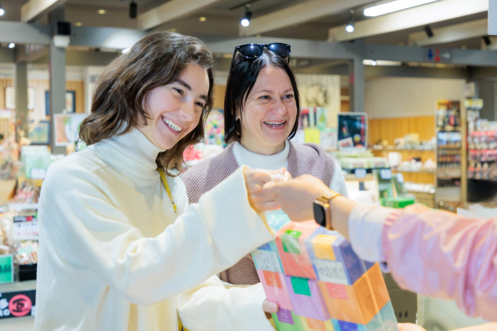 Two smiling shoppers hand a colorful boxed gift to a staff member across the counter of a Japanese souvenir shop