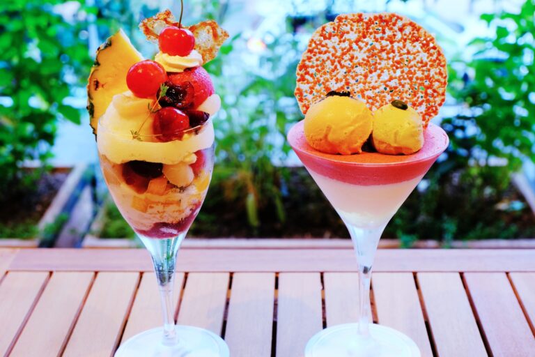 Two parfait glasses on a wooden table, one topped with pineapple and cherries, the other with mango scoops on pink mousse.