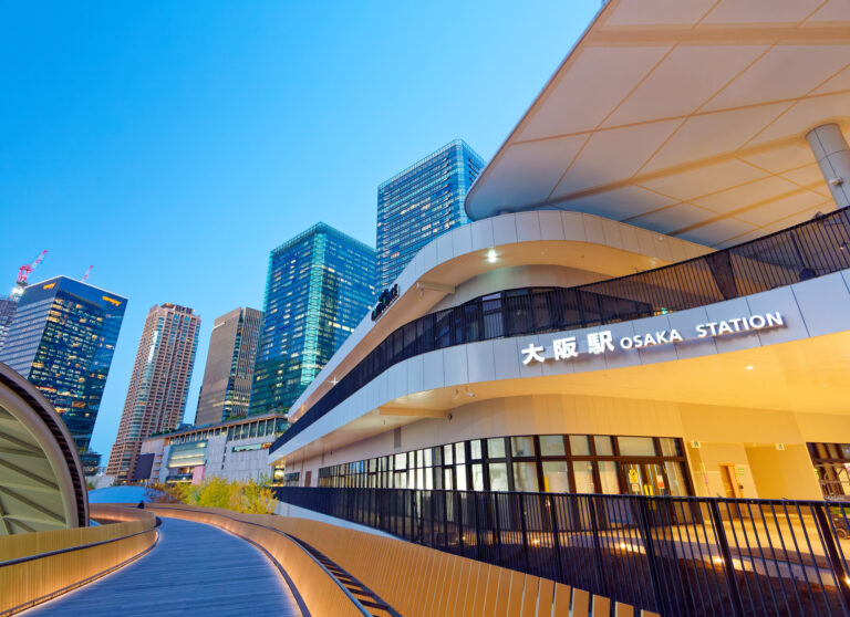 JR Osaka Station's illuminated modern exterior at dusk, with a curving elevated walkway and glass office towers behind.