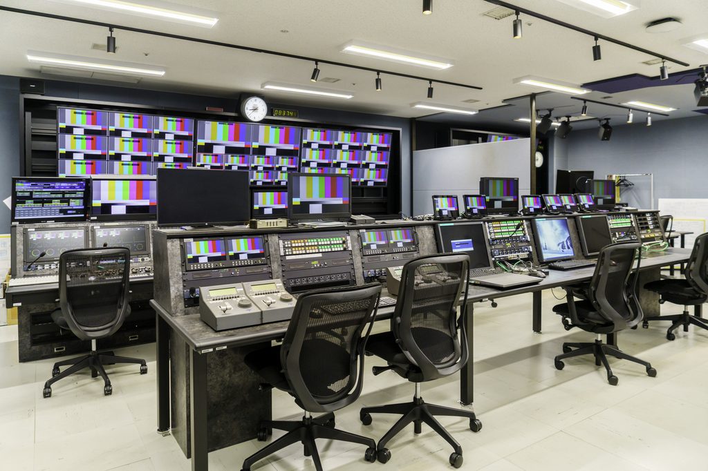 A television station's master control room, banks of monitors showing color test bars above an empty mixing desk.