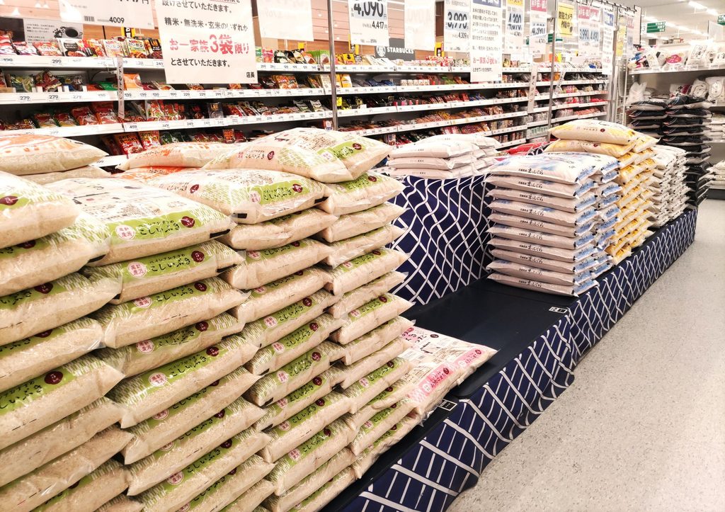 Stacked 5kg bags of rice in a supermarket beneath price tags and a sign limiting purchases to three bags per family.