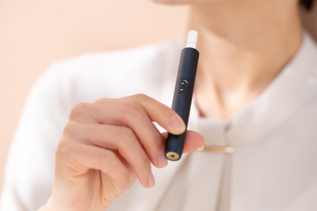 Close-up of a person in a white blouse holding a black heated-tobacco device loaded with a white tobacco stick.