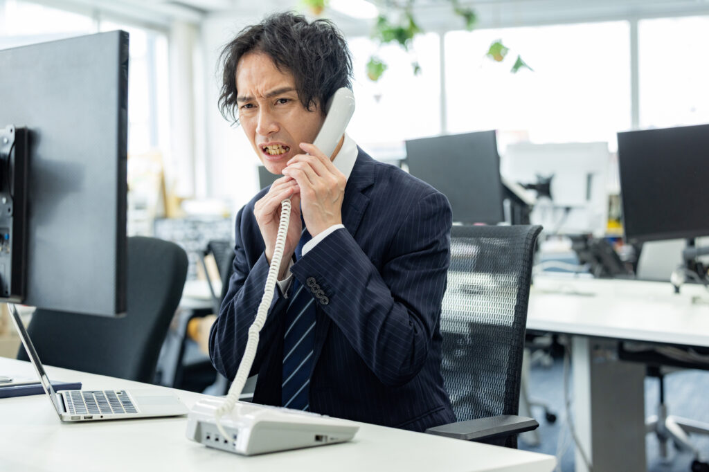 A businessman grips a landline phone with a tense, frowning expression at his desk
