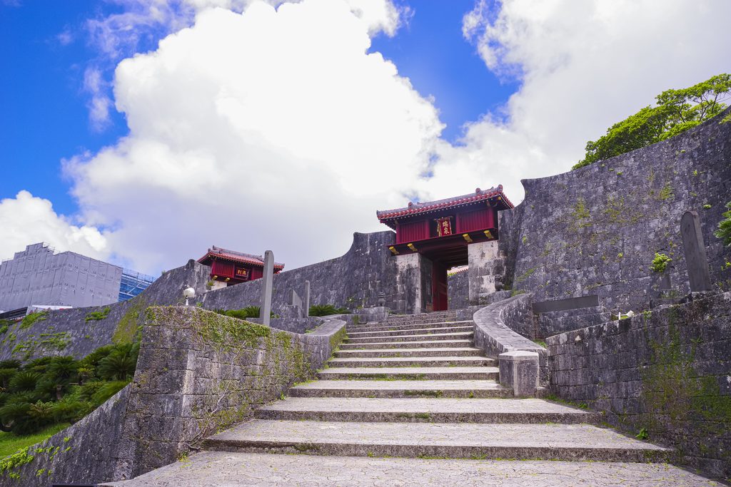 Stone stairs climbing between limestone walls toward a red-roofed gate at Shuri Castle, under a bright cloudy sky