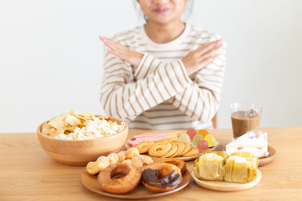 A woman crosses her arms in an X gesture in front of a table full of chips, donuts, and other snacks.