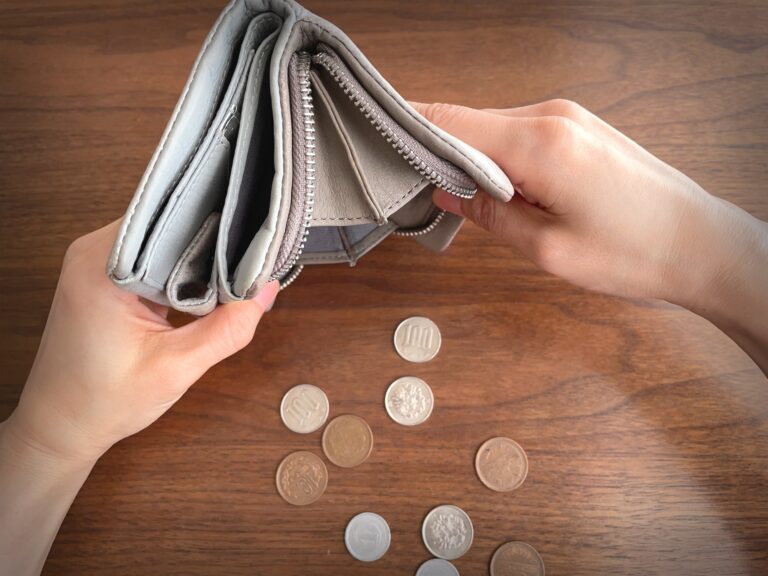 Two hands hold an empty grey wallet open over a wooden table, with Japanese 100, 10, 5 and 1 yen coins scattered beside it.