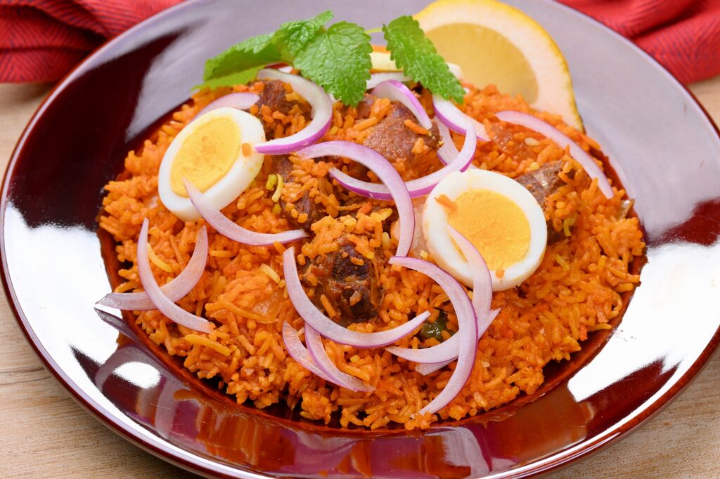 Mutton biryani with halved boiled eggs, sliced red onion and a lemon wedge on a dark red plate.