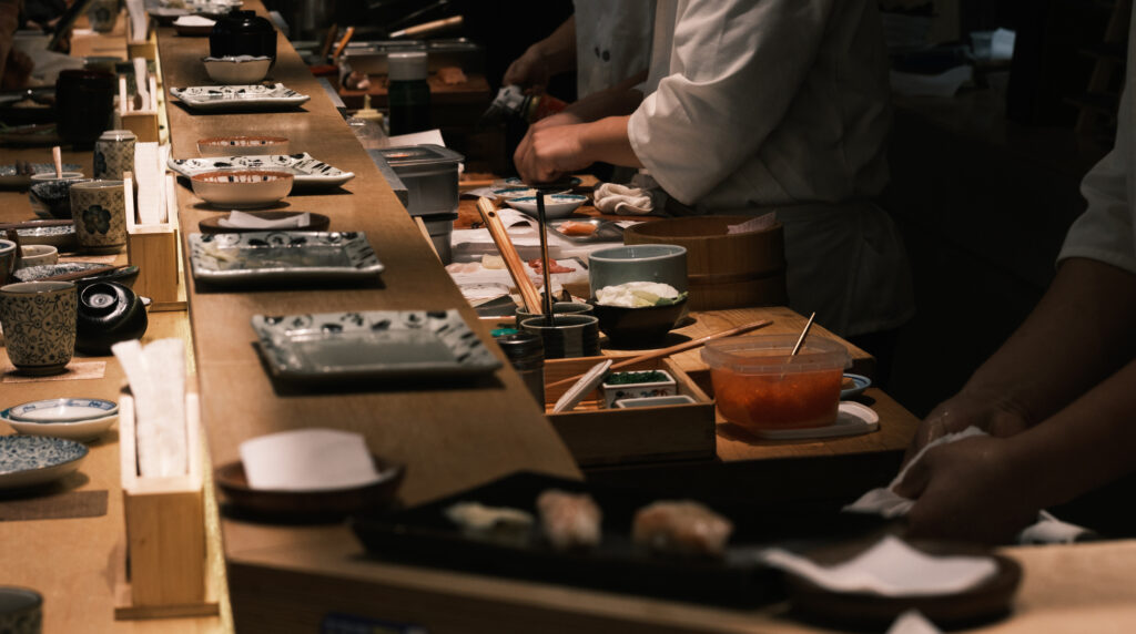 A sushi chef prepares nigiri behind a traditional wooden counter lined with ceramic plates and condiments.