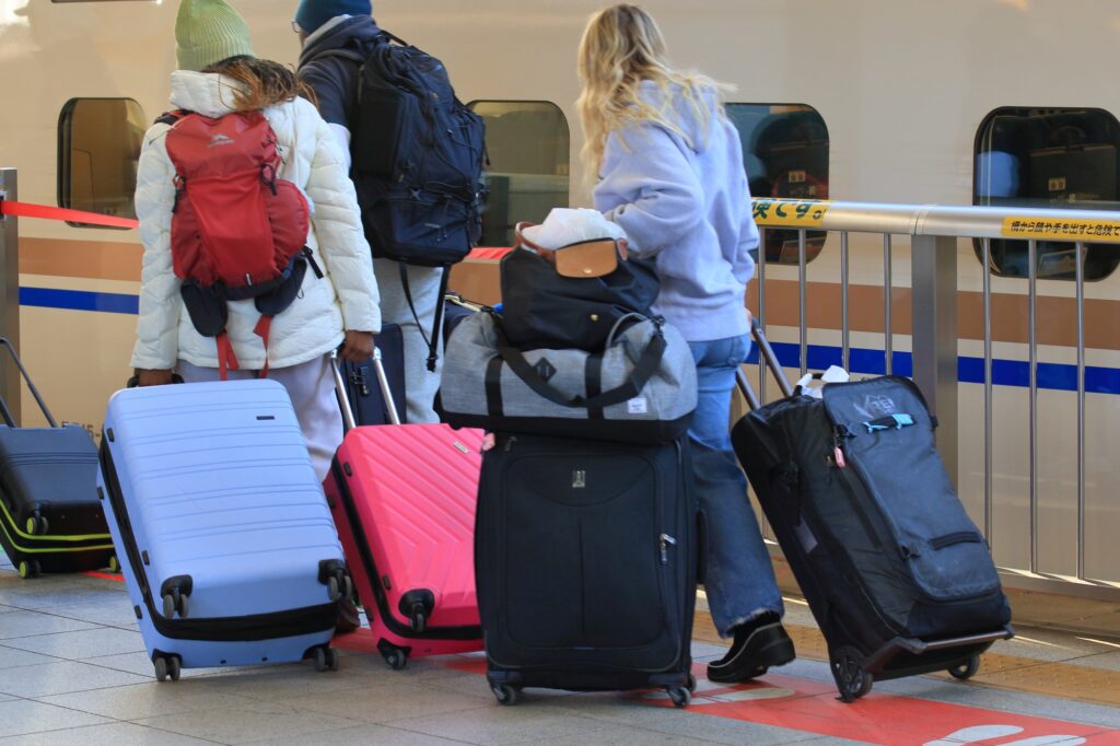 Three travelers seen from behind on a Shinkansen platform with five large rolling suitcases and backpacks.