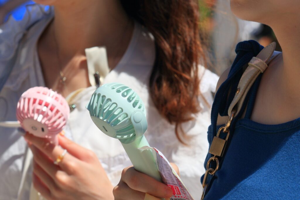Two people in a sunlit crowd hold pink and mint-green handheld electric fans up toward their faces.