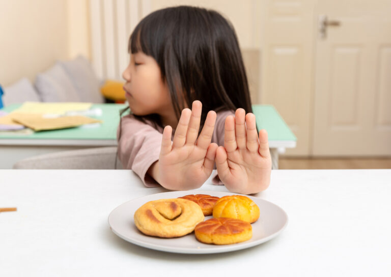 A young girl holds up both hands to refuse a plate of pastries on a table.