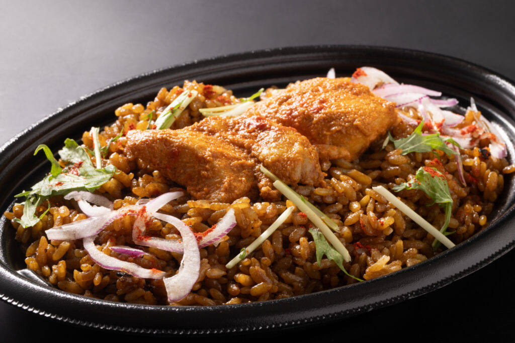 Chicken biryani in a black oval dish, topped with sliced red onion, herbs and chili powder.