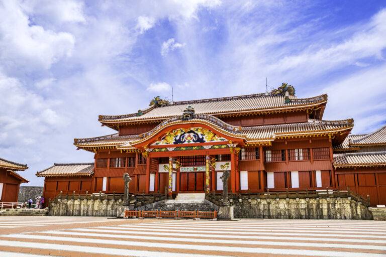 Shuri Castle's red main hall seen across its stone plaza, with dragon roof ornaments and stone pillars flanking the stairs