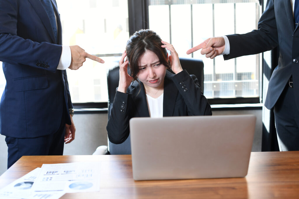 Two men in suits point at a woman sitting at a laptop between them in an office