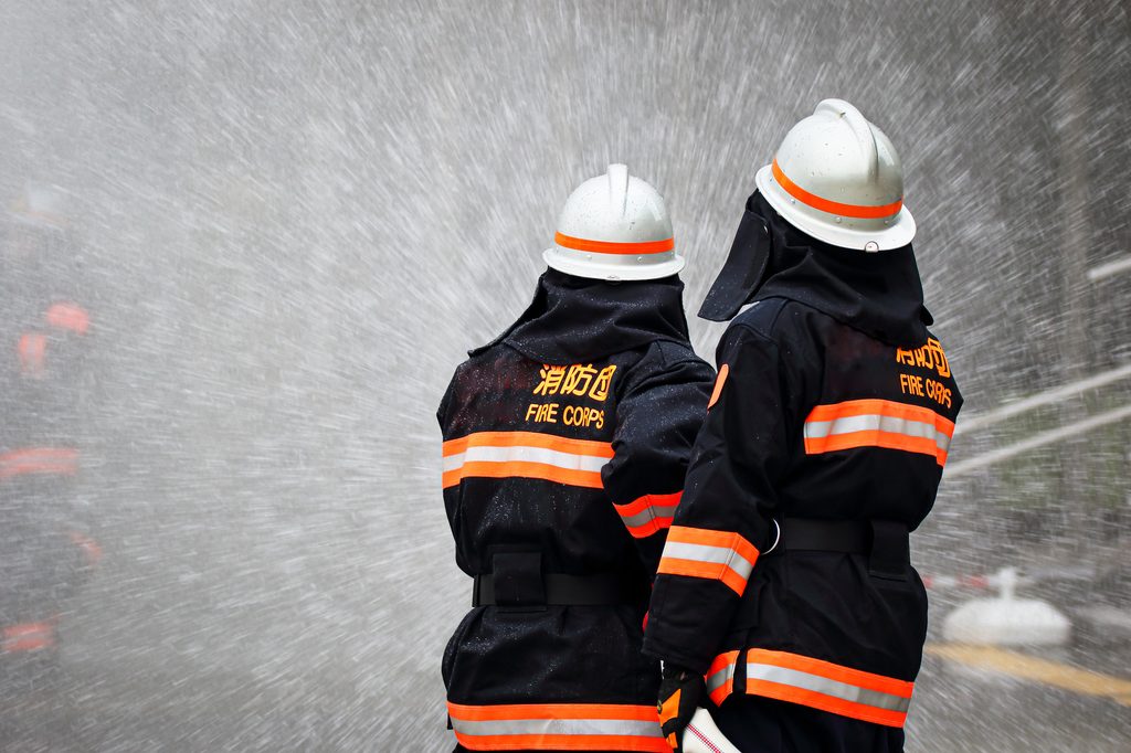 Two Japanese firefighters seen from behind in black turnout gear reading Fire Corps, standing in heavy water spray