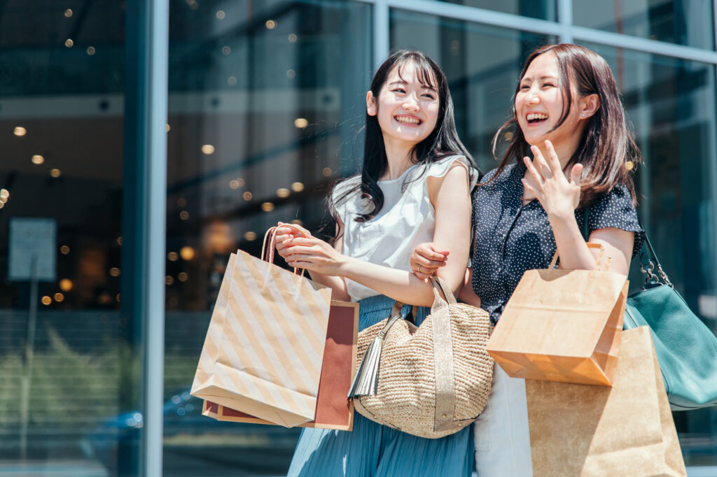 Two young women laugh outside a glass-fronted store, carrying paper shopping bags and a woven straw tote