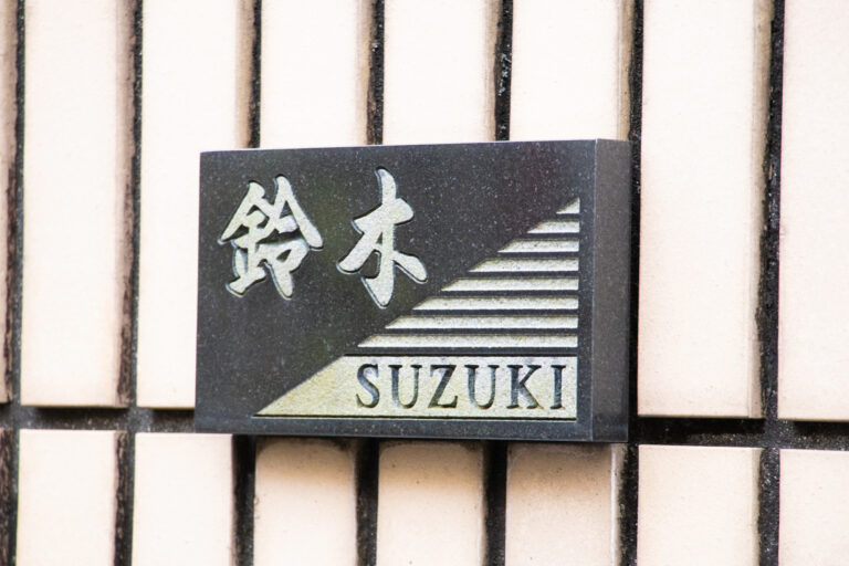 A stone nameplate reading 鈴木 and SUZUKI mounted on the tiled exterior wall of a Japanese house.