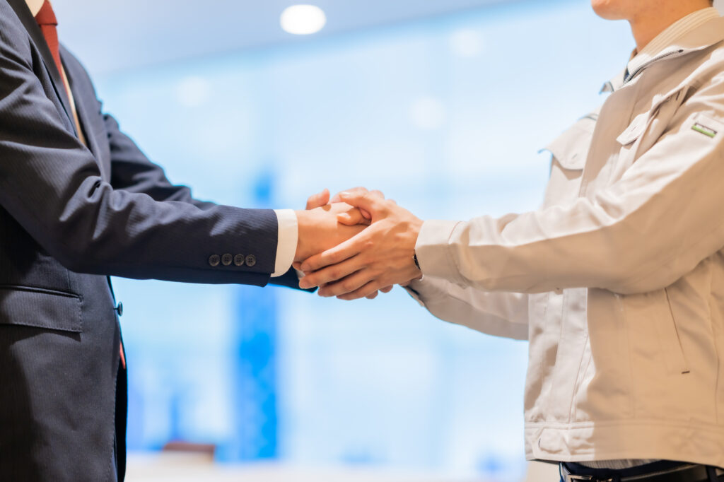 A businessman in a suit shakes hands with a worker wearing a beige work jacket