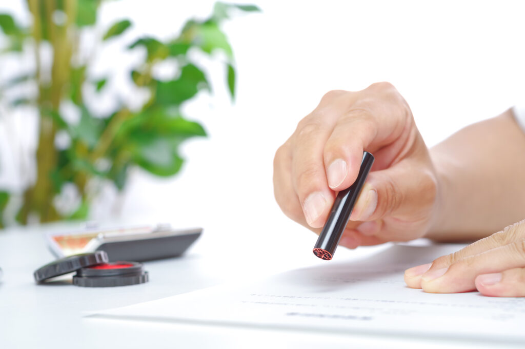 A hand pressing a personal name seal onto a printed Japanese document, with a red ink pad nearby.