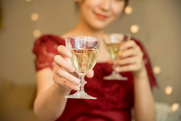 Woman in a red dress holds a glass of sparkling wine toward the camera, a second glass raised behind her.