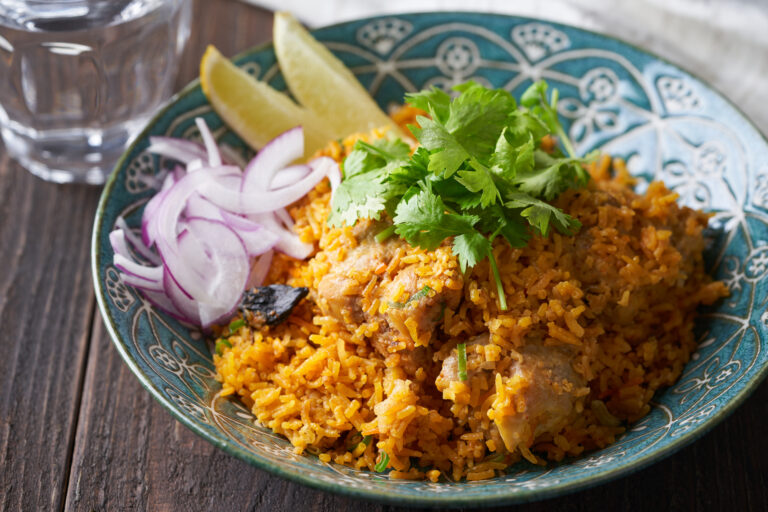 Chicken biryani on a patterned blue-green plate with cilantro, sliced red onion and lemon wedges, on a dark wood table.