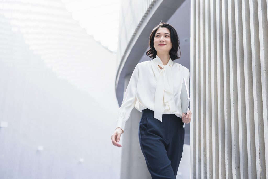 Woman in a white blouse and navy trousers walks outdoors carrying a laptop.