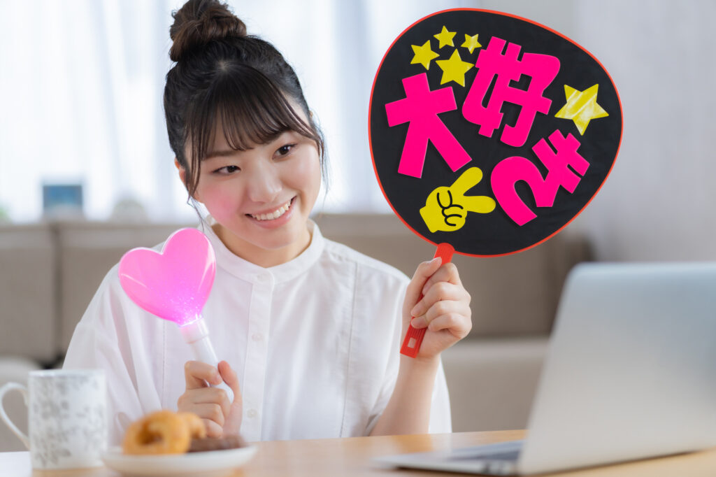 A young woman at her laptop holds a heart-shaped light stick and a fan sign, with a donut on a plate beside her.