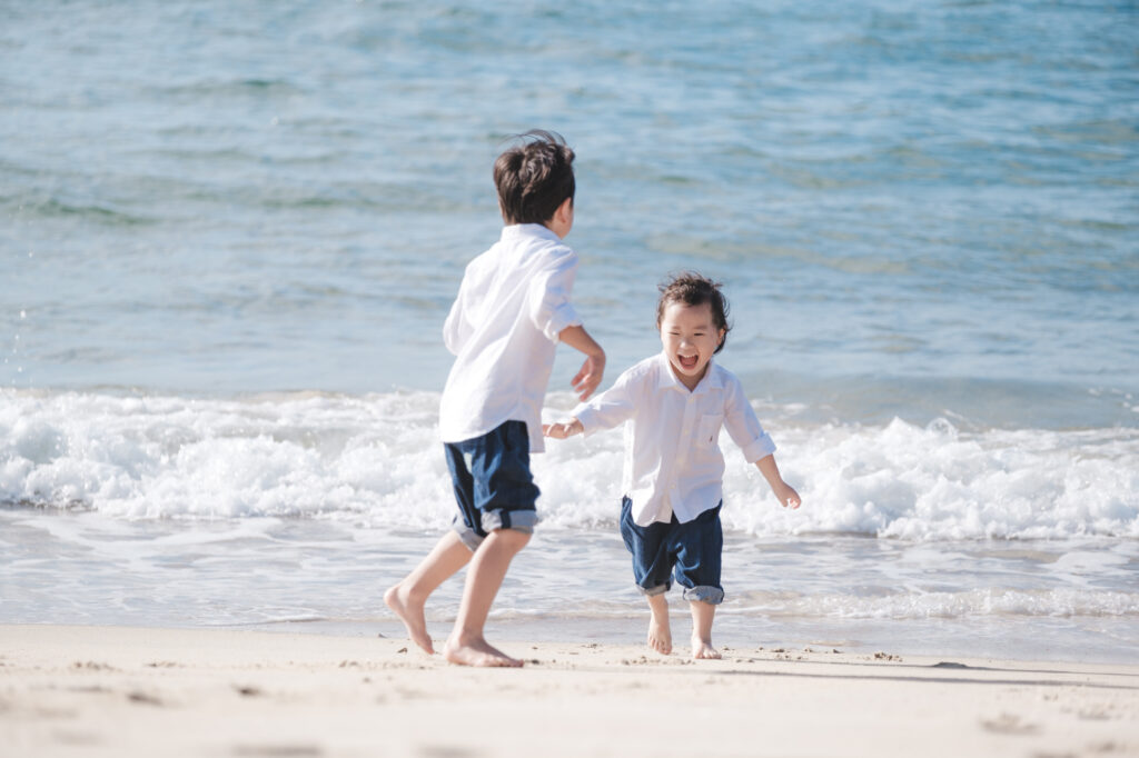 Two young children in white shirts and rolled denim shorts run barefoot along the wet sand at the water's edge.