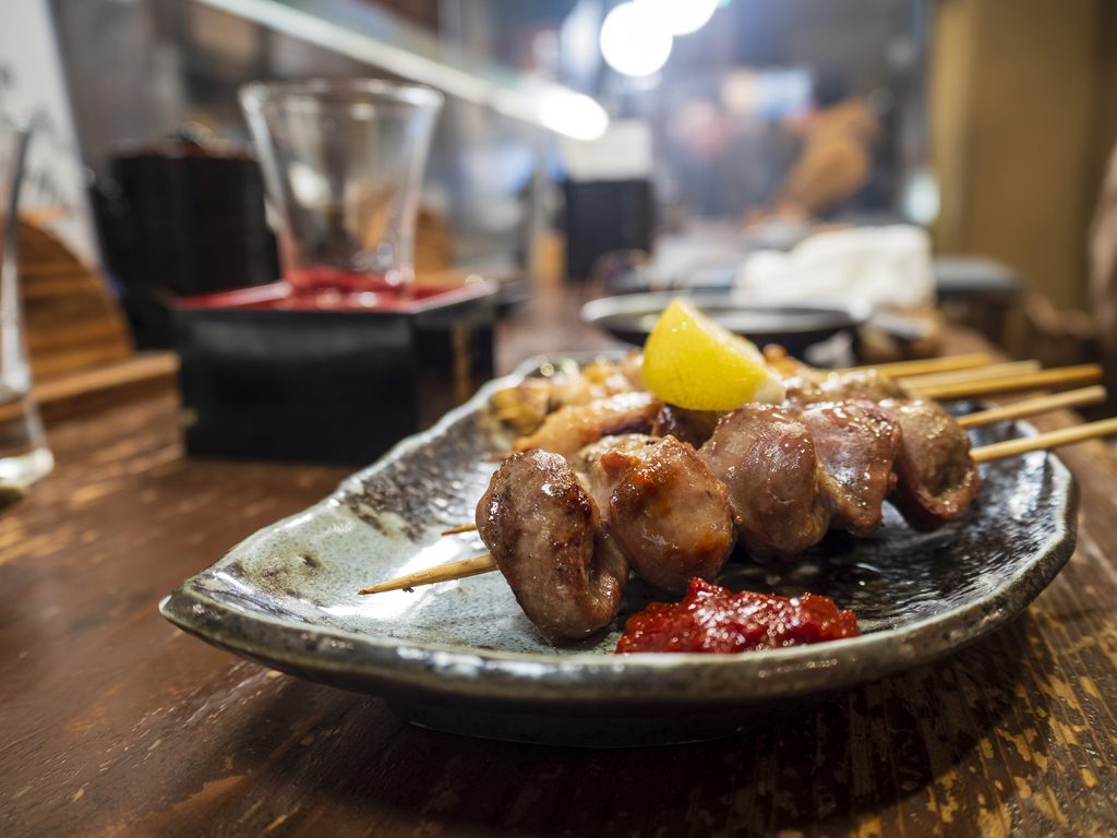 Grilled chicken skewers on a ceramic plate with a lemon wedge and red chili paste, on a wooden counter.