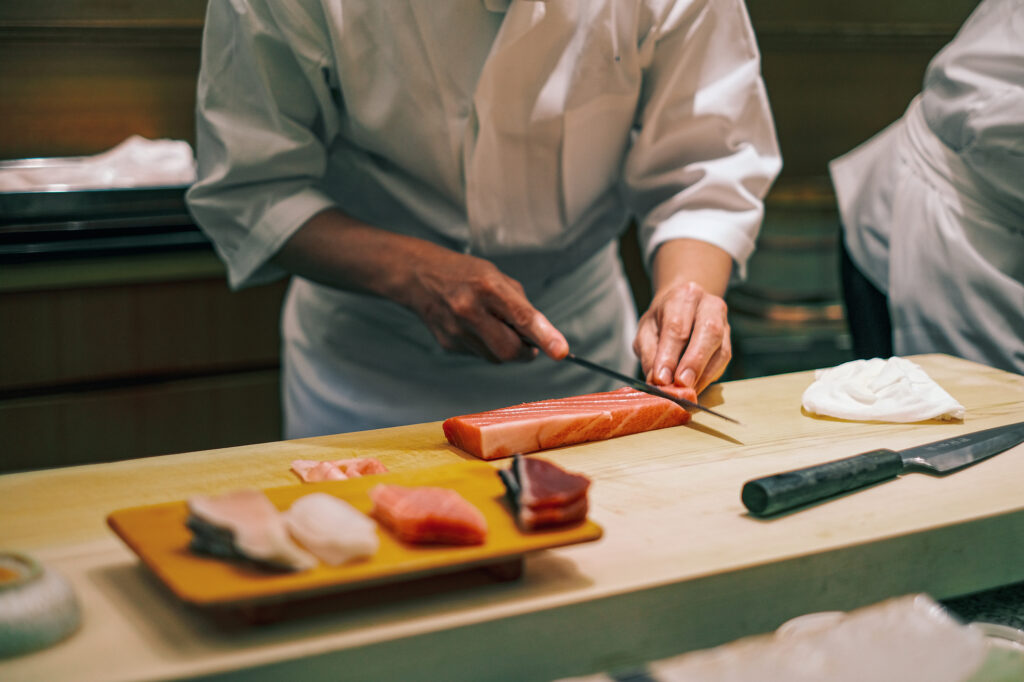 Sushi chef in white slicing a block of tuna with a long knife at a counter, with cut fish on a board nearby.