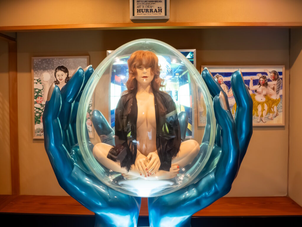 A mannequin of a woman seated inside a clear egg-shaped capsule held by two giant blue hands, framed artworks behind