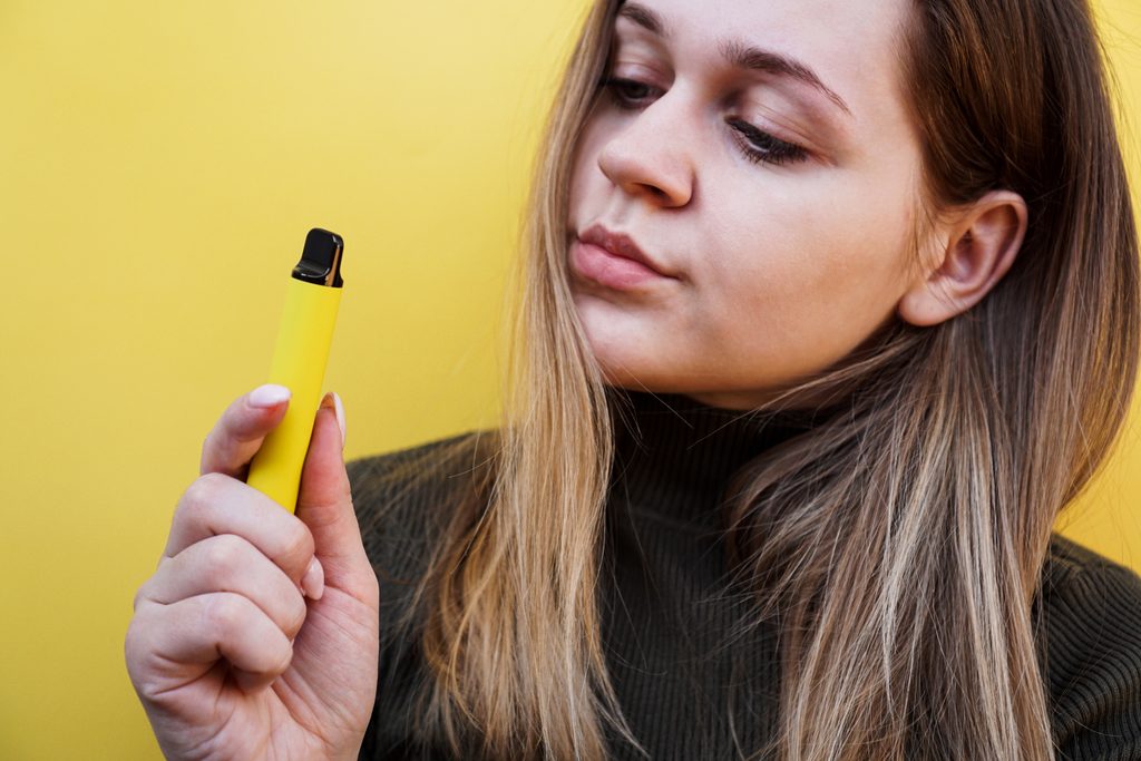 A young woman with long blonde hair examines a yellow disposable vape she is holding, against a yellow background.