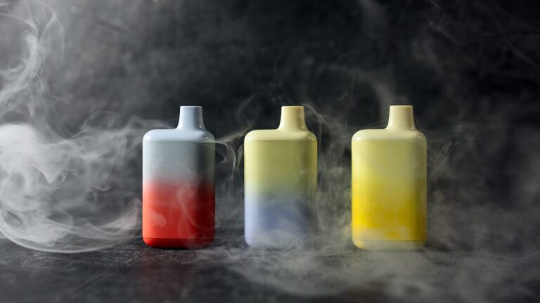 Three brightly colored disposable vape devices standing on a dark surface, surrounded by drifting vapor.