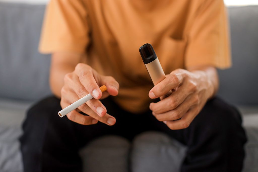 A person seated on a sofa holds a cigarette in one hand and a gold disposable vape in the other.