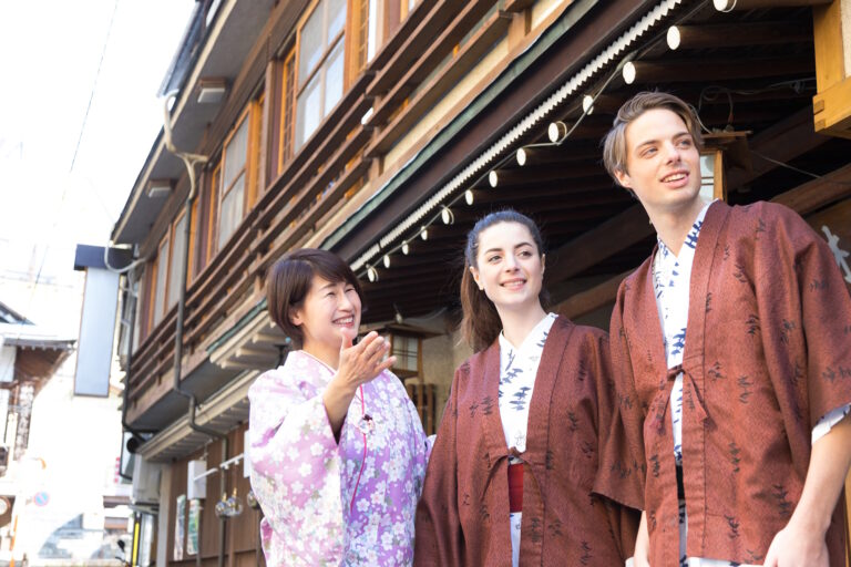 A guide in a floral kimono points something out to a Western couple in haori on a traditional shopping street