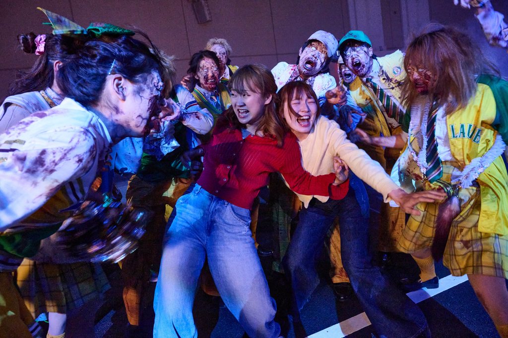 A crowd of costumed zombies in torn school uniforms lurches across a floodlit park street at night