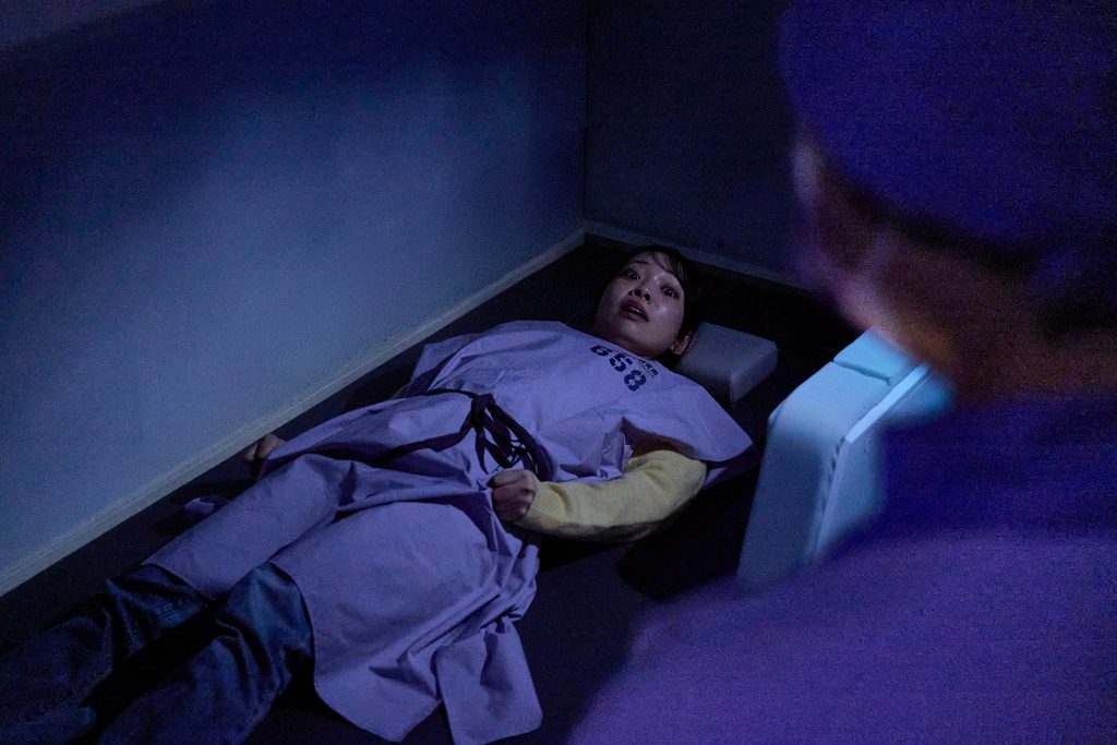 A guest in a numbered gown lies on the floor of a bare blue-lit cell, watched by a figure in a surgical cap