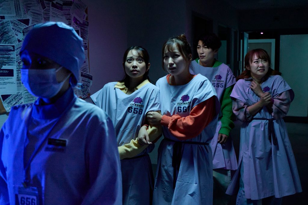 Four guests in numbered hospital gowns huddle in a dim corridor as a masked figure in a lab coat passes